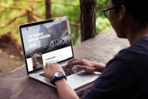 A person sitting at a wooden table outdoors, using a laptop. The screen displays a website with an RV and the text Live the Great American Adventure. The person is wearing glasses and a watch, with a blurred green background.